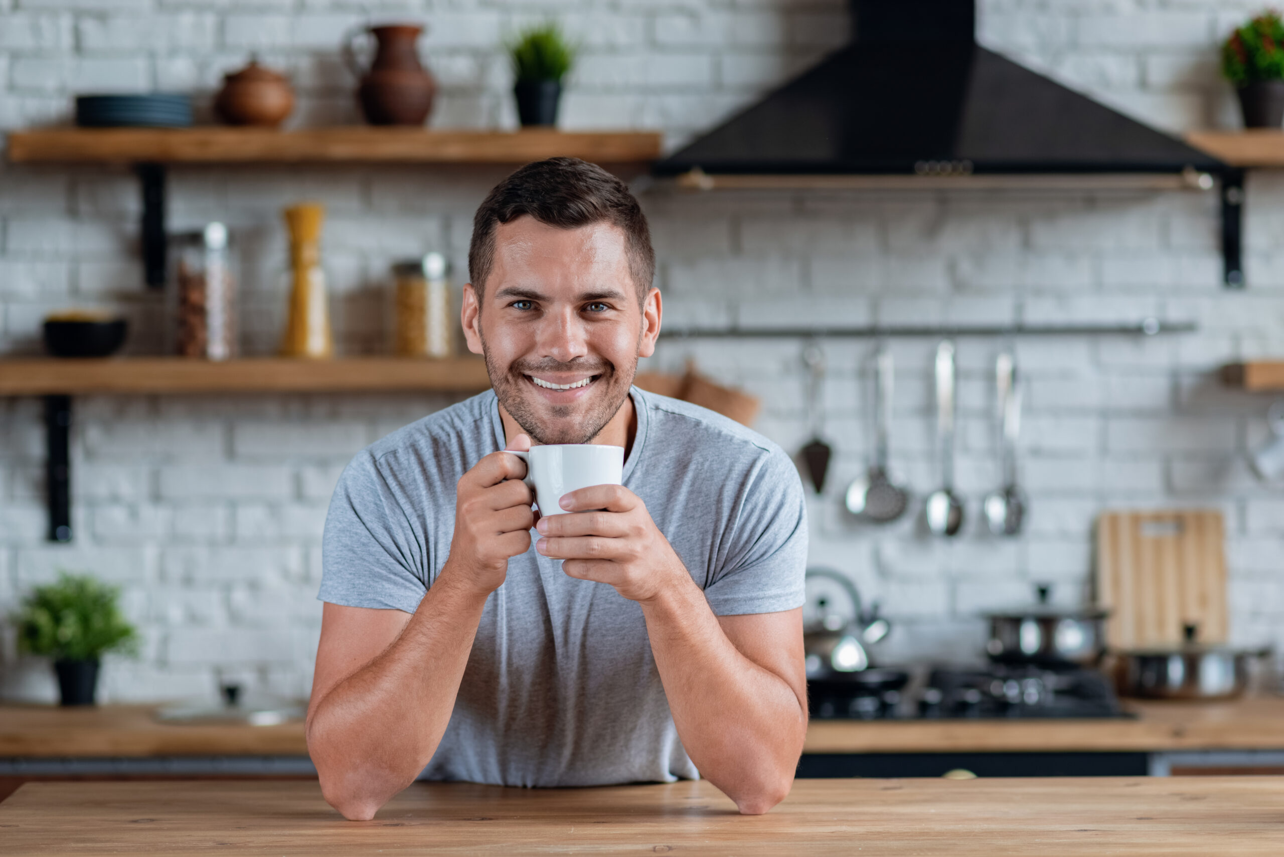 Handsome Man Sits At The Kitchen Table With Cup Of Morning Tea Or Coffee And Smiling Looking At The Camera