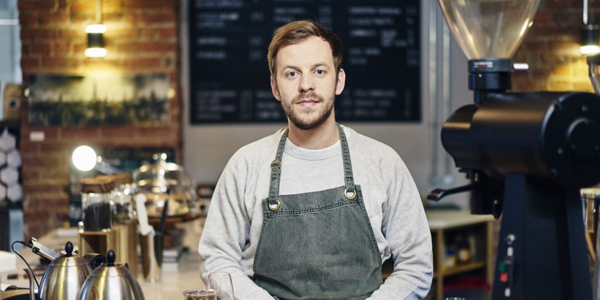 Portrait Of Young Male Barista At Coffee Shop Kitchen Counter